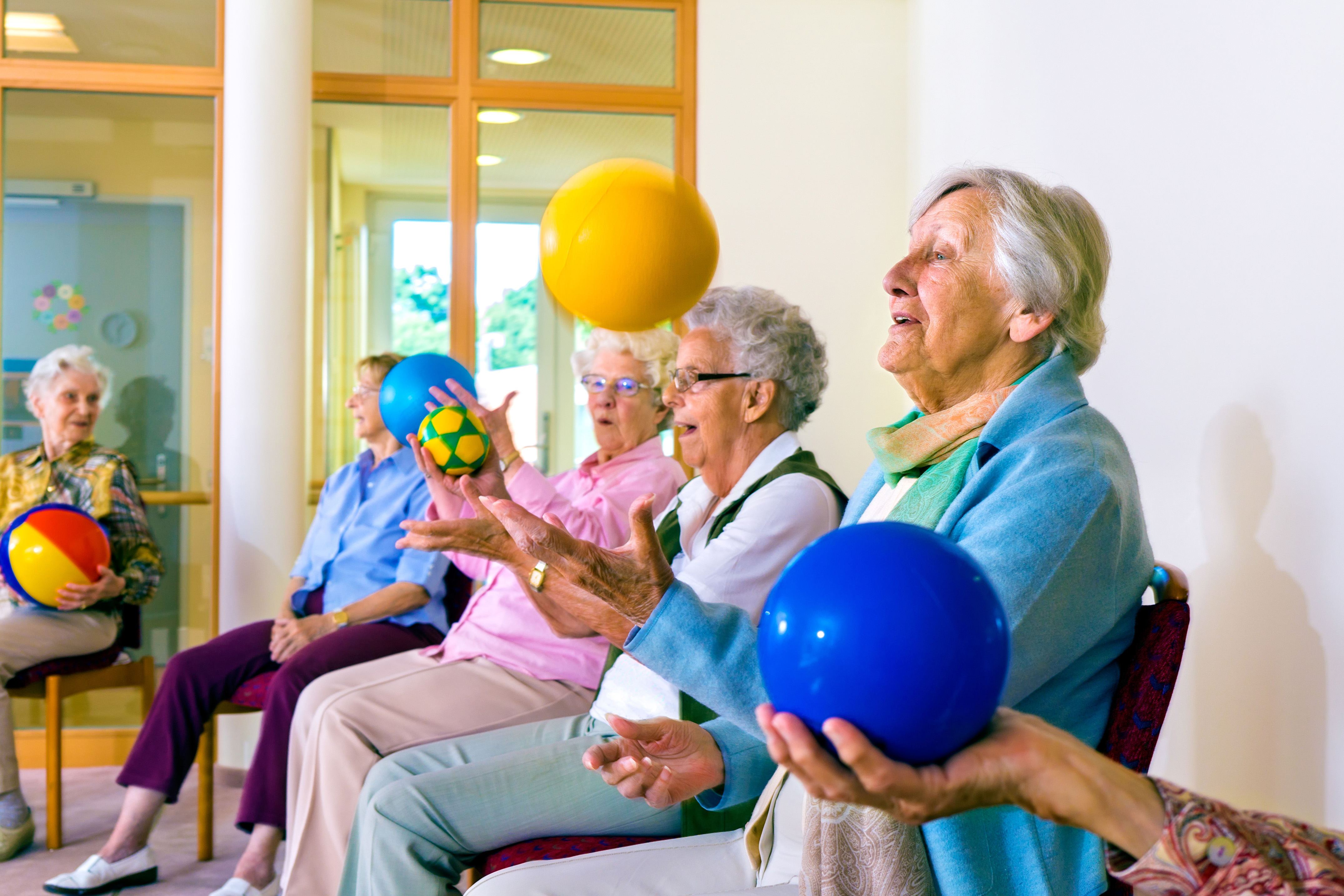 Group of happy senior ladies doing coordination exercises in a seniors gym sitting in chairs throwing and catching brightly colored balls